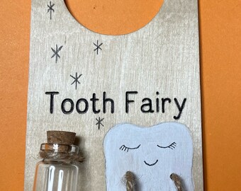 May include: A wooden door hanger with a tooth fairy theme. It features a small glass jar with a cork stopper, a white tooth with a smiling face, and the text "Tooth Fairy" and "Please Stop Here".