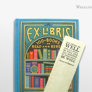 May include: A gold bookmark with the text "All shall be well and all shall be well and all manner of thing shall be well" by Julian of Norwich. The bookmark is on top of a blue book with the text "Ex-Libris 100+ Books to Read and Rere" and a picture of a bookshelf.