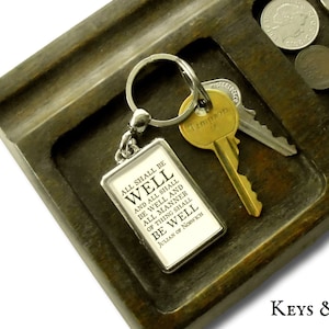 May include: A silver keychain with a rectangular charm that reads "All shall be well and all shall be well and all manner of thing shall be well Julian of Norwich". The keychain is on a wooden tray with coins.