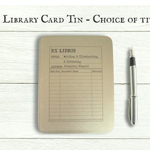 May include: A gold-coloured metal tin with a library card design. The tin has the text "EX LIBRIS" and "Writing & Illuminating & Lettering" printed on it. The tin also has a section for the author, date due, borrower's name, and returned.