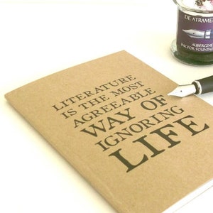 Puede incluir: Una libreta marr&oacute;n con el texto "LITERATURE IS THE MOST AGREEABLE WAY OF IGNORING LIFE" impreso en la portada. Tambi&eacute;n se ve una pluma plateada y un peque&ntilde;o frasco de tinta en la imagen.
