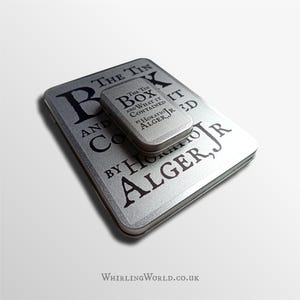May include: Two silver rectangular tin boxes, one larger than the other, are stacked. The text "THE TIN BOX AND WHAT IT CONTAINED" and "BY HORATIO ALGER, JR" is printed in black on the boxes. The website "WhirlingWorld.co.uk" is at the bottom.