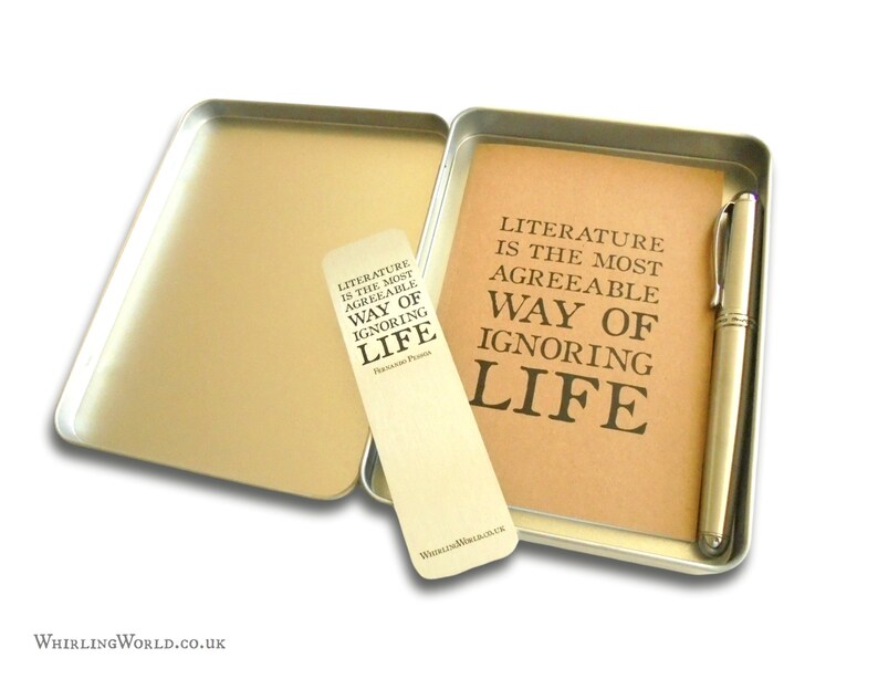 May include: A silver metal tin with a brown paper insert and a silver pen. The paper insert has the text "Literature is the most agreeable way of ignoring life" printed on it. The tin also includes a bookmark with the same text printed on it.