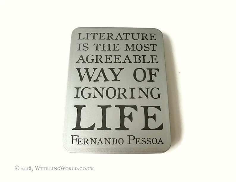 May include: A silver metal sign with the quote "Literature is the most agreeable way of ignoring life" by Fernando Pessoa.