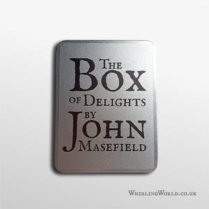 May include: A silver metal plaque with rounded corners, featuring the text "The Box of Delights by John Masefield" in a dark serif font. The website "WhirlingWorld.co.uk" is displayed at the bottom of the plaque.