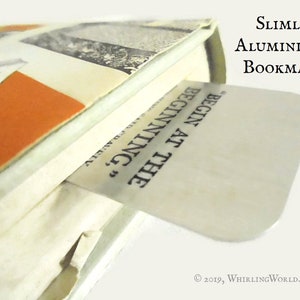 May include: A silver bookmark with the text "BEGIN AT THE BEGINNING" printed on it. The bookmark is made of slimline aluminum.