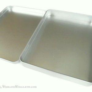 May include: A silver metal box with a hinged lid. The box is empty and has a smooth, shiny surface.