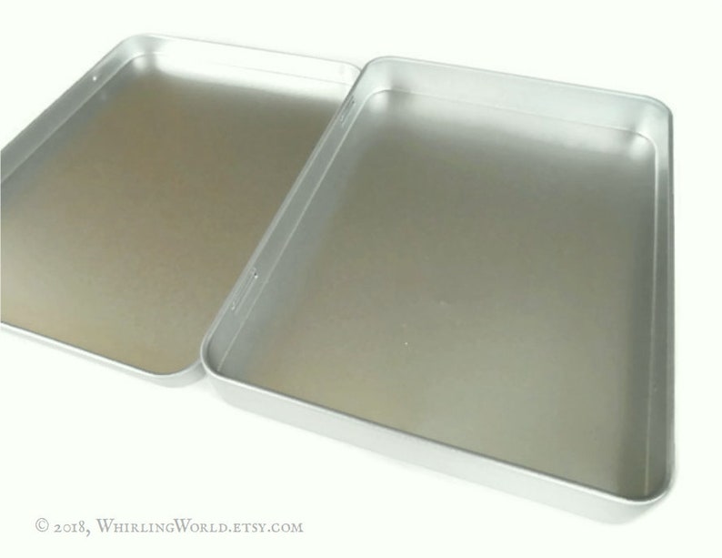 May include: A silver metal box with a hinged lid. The box is empty and has a smooth, shiny surface.