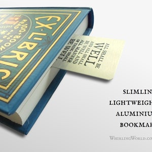 May include: A silver bookmark with the text "All shall be well and all shall be well and all manner of thing shall be well" is tucked into the corner of a blue book with gold lettering that reads "Ex Libris". The bookmark is described as "Slimline, Lightweight Aluminium Bookmark" and the website "WhirlingWorld.co.uk" is printed below.