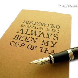 May include: A brown notebook with the text "Distorted realities have always been my cup of tea" printed on the cover. A black and silver fountain pen is lying on top of the notebook.