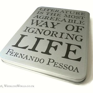 May include: A silver metal box with the quote "Literature is the most agreeable way of ignoring life" by Fernando Pessoa printed on the front.