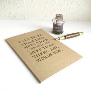 May include: A tan notebook with the quote "I SEE MORE THAN THIS, MORE THAN I CAN TELL YOU, MORE THAN THERE ARE WORDS FOR." is next to a fountain pen and a bottle of ink.