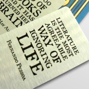May include: A silver bookmark with a quote about literature being the most agreeable way of ignoring life. The quote is attributed to Fernando Pessoa.