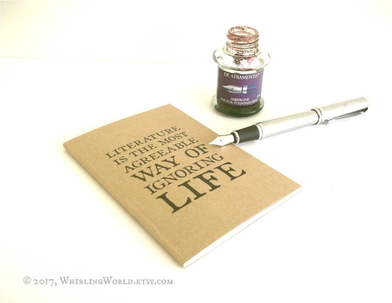 Puede incluir: Una libreta marr&oacute;n con el texto "LITERATURE IS THE MOST AGREEABLE WAY OF IGNORING LIFE" impreso en la portada. Tambi&eacute;n se ve una pluma plateada y un peque&ntilde;o frasco de tinta en la imagen.