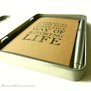 Puede incluir: Una caja de metal plateada con un cuaderno marr&oacute;n y un bol&iacute;grafo plateado en su interior. El cuaderno tiene el texto "Literature is the most agreeable way of ignoring life" impreso en &eacute;l.