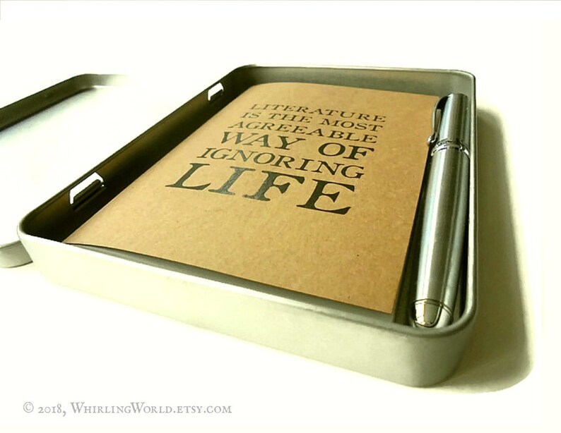 Puede incluir: Una caja de metal plateada con un cuaderno marr&oacute;n y un bol&iacute;grafo plateado en su interior. El cuaderno tiene el texto "Literature is the most agreeable way of ignoring life" impreso en &eacute;l.