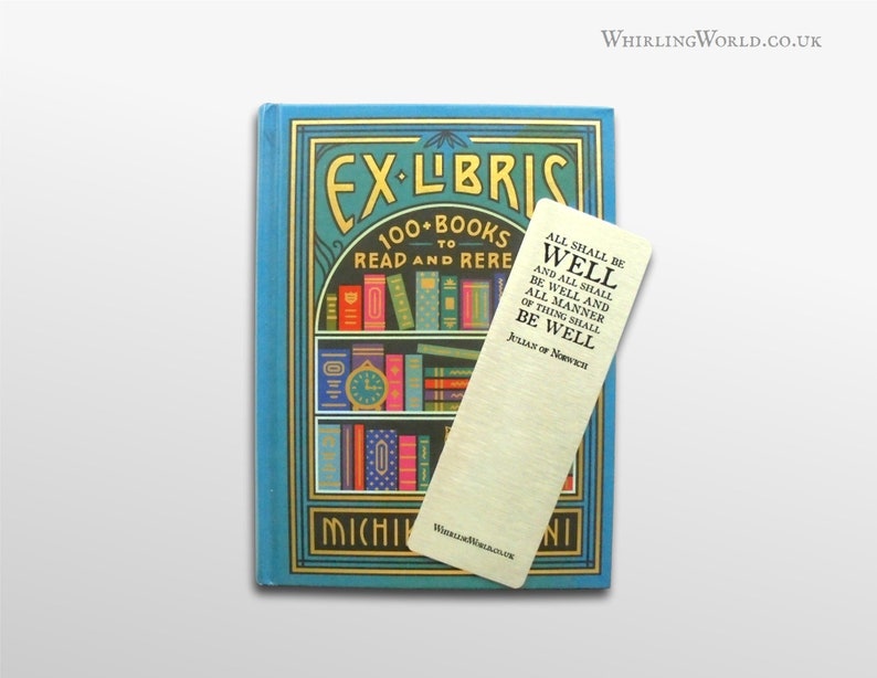 May include: A gold bookmark with the text "All shall be well and all shall be well and all manner of thing shall be well" by Julian of Norwich. The bookmark is on top of a blue book with the text "Ex-Libris 100+ Books to Read and Rere" and a picture of a bookshelf.