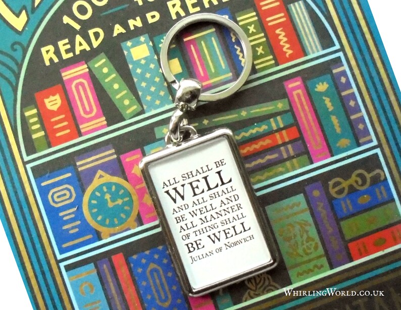 May include: A silver keychain with a small rectangular tag that reads "All shall be well and all shall be well and all manner of thing shall be well Julian of Norwich". The keychain is in front of a book with a colorful bookshelf design.