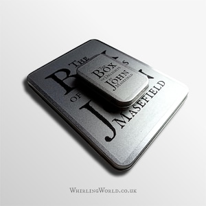 May include: Two silver metal tins are shown, one rectangular and one smaller. The larger tin features the text "THE BOX OF DELIGHTS BY JOHN MASEFIELD" in black. The smaller tin has the same text. The website "WhirlingWorld.co.uk" is printed at the bottom.