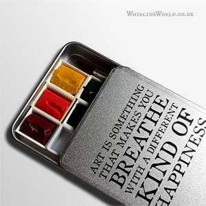 May include: A silver watercolour paint set with a hinged lid. The open lid displays the text "ART IS SOMETHING THAT MAKES YOU BREATHE WITH A DIFFERENT KIND OF HAPPINESS." The paint set contains various colours, including yellow, red, and dark red.