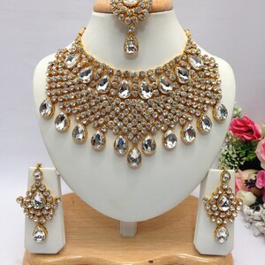 May include: A gold-tone bridal necklace set with clear teardrop-shaped crystals and smaller round crystals. The set includes a necklace, earrings, and a maang tikka.