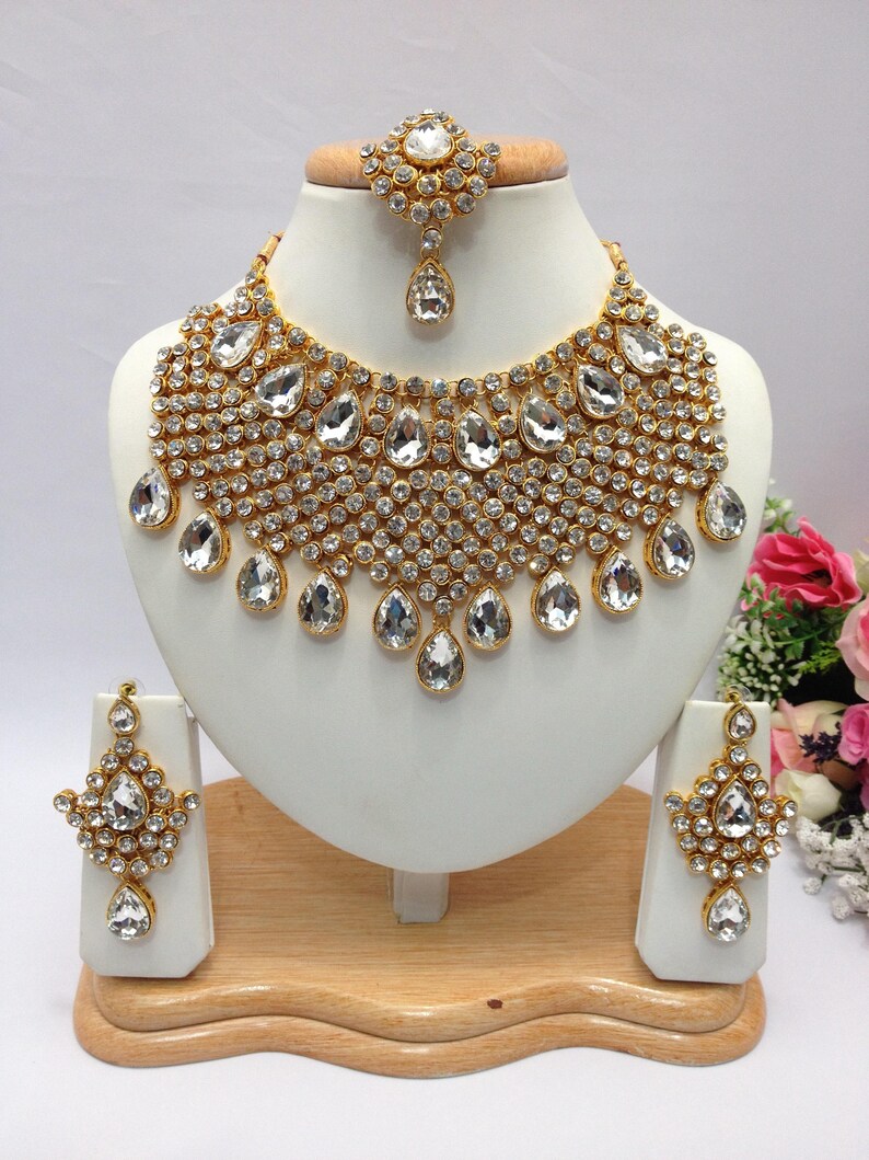 May include: A gold-tone bridal necklace set with clear teardrop-shaped crystals and smaller round crystals. The set includes a necklace, earrings, and a maang tikka.