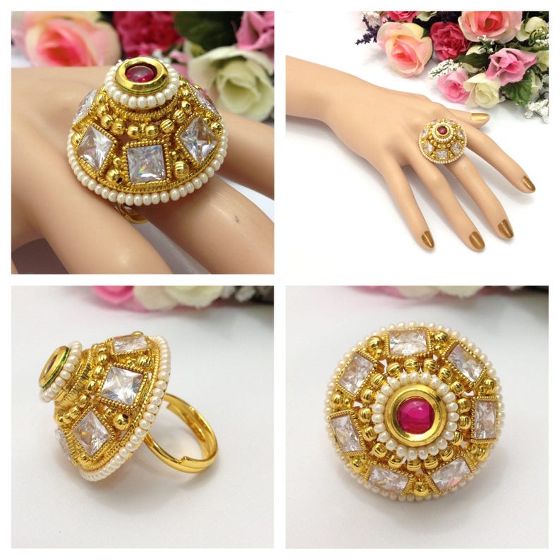 May include: A gold ring with a round, dome-shaped design. The ring is adorned with white cubic zirconia stones and a single pink stone in the centre. The ring is surrounded by a row of small white pearls.