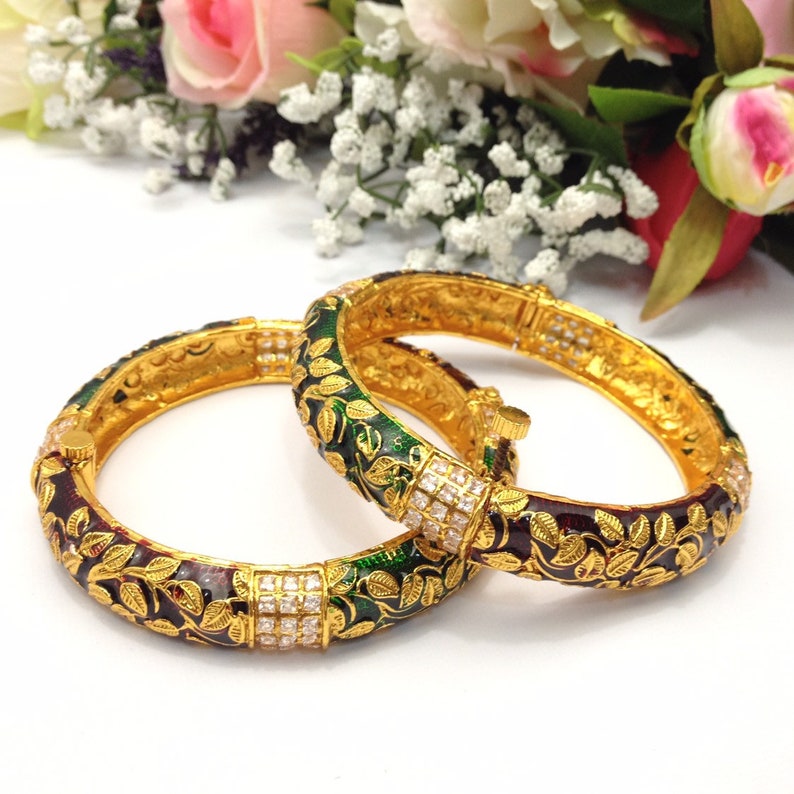 May include: Two gold-colored bangles with intricate leaf and vine designs. The bangles feature a combination of dark red and green enamel, accented with small, square-cut clear stones. The background includes pink and white flowers.
