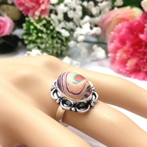 Può includere: Un anello d'argento con una grande pietra ovale. La pietra è un mix di colori rosa, viola, arancio e turchese. L'anello ha un design floreale decorativo attorno alla pietra.