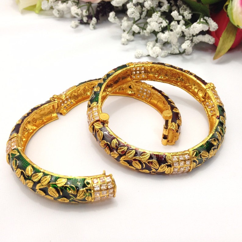 May include: Two gold-toned bangles with intricate leaf and floral designs. The bangles feature green, burgundy, and gold enamel, accented with square-cut clear stones. The open design allows for easy wear.