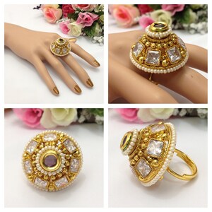 May include: A gold ring with a large, round, white stone centre surrounded by smaller white stones and a single purple stone. The ring is decorated with small white pearls.