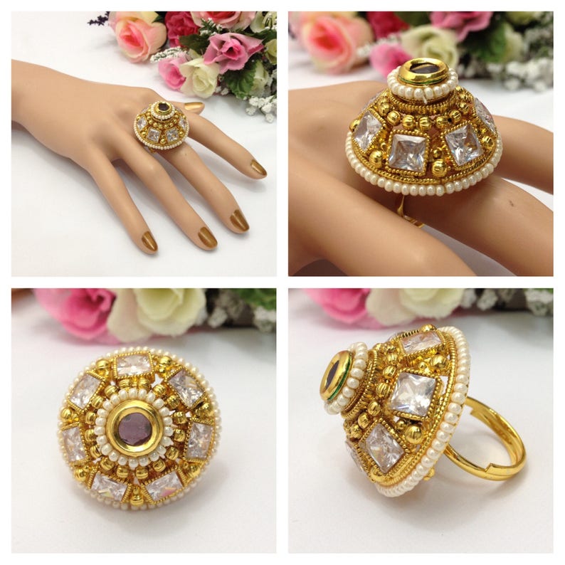 May include: A gold ring with a large, round, white stone centre surrounded by smaller white stones and a single purple stone. The ring is decorated with small white pearls.