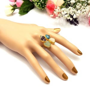 May include: A gold-toned ring featuring five irregularly shaped gemstones in shades of purple, blue, white, and green. The ring is set on a mannequin hand with gold-colored fingernails, against a backdrop of flowers.