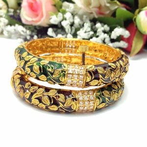 May include: Two gold-toned bangles with intricate leaf designs in green and burgundy enamel. The bangles feature a square, gem-studded clasp and are stacked on top of each other. The background includes flowers.