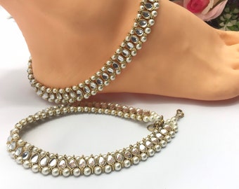 May include: Gold-toned anklets with pear-shaped clear stones and small white pearls. The anklets are displayed on a foot and against a white background. The jewelry has a delicate, ornate design.