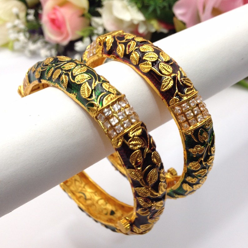 May include: Two gold bangles with intricate leaf and vine designs. The bangles feature a dark green and burgundy enamel background with gold leaf accents. A section of the bangles is adorned with square-cut, clear gemstones.