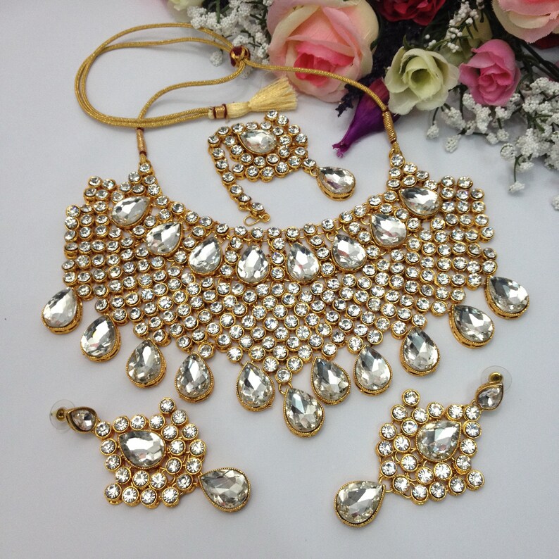 May include: A gold-tone bridal necklace set with clear teardrop-shaped crystals and smaller round crystals. The set includes a necklace, earrings, and a tikka.