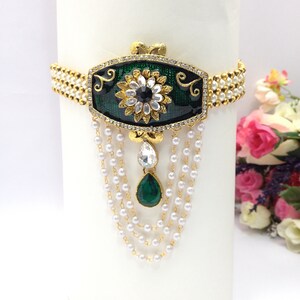 Op de afbeelding: Een gouden ketting met een groene en zwarte emaille hanger met een witte bloem met een zwart hart. De hanger is omringd door heldere strass steentjes en hangt aan een ketting van witte parels.