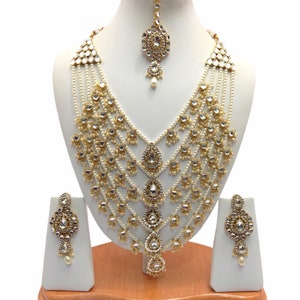 Puede incluir: Un collar de cinco hileras de oro y perlas blancas con un colgante en forma de lágrima. El collar está adornado con piedras transparentes y blancas. El conjunto también incluye pendientes a juego y un tikka.