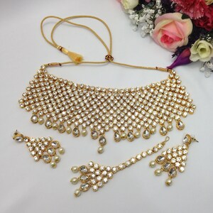 May include: A gold and white Indian bridal necklace set featuring a choker style necklace, matching earrings, and a maang tikka. The set is decorated with clear stones and pearls.