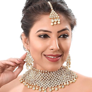 May include: A gold and white bridal necklace set featuring a large, ornate choker, matching earrings, and a traditional forehead ornament. The set is decorated with sparkling white stones and pearls.