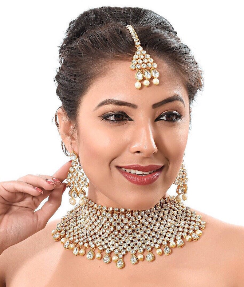 May include: A gold and white bridal necklace set featuring a large, ornate choker, matching earrings, and a traditional forehead ornament. The set is decorated with sparkling white stones and pearls.