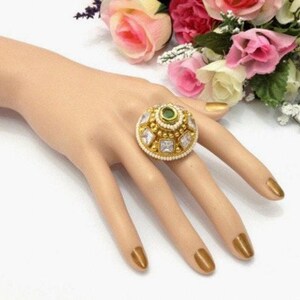 May include: A gold ring featuring a large, round, green gemstone encircled by smaller, clear gemstones. The ring is adorned with white pearls around the edge.