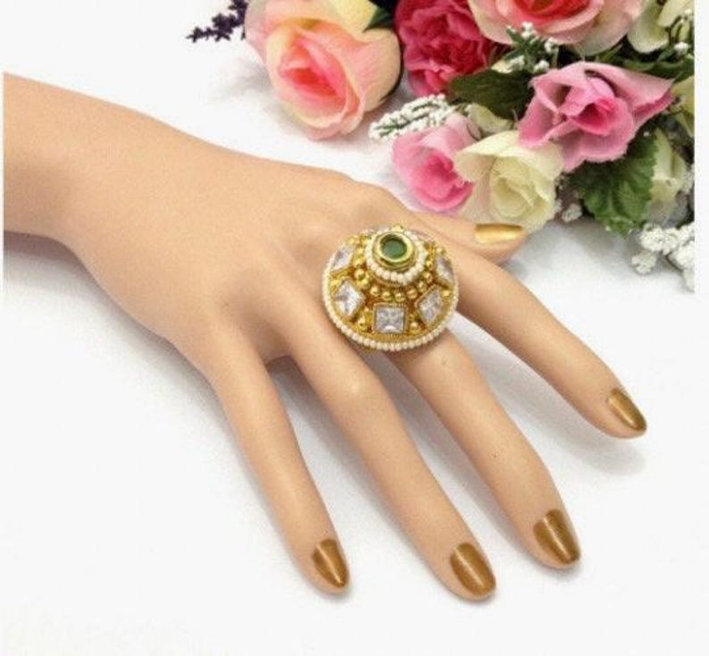 May include: A gold ring featuring a large, round, green gemstone encircled by smaller, clear gemstones. The ring is adorned with white pearls around the edge.