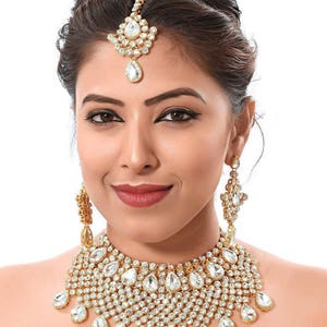 May include: A gold and white bridal jewellery set with a necklace, earrings, and a tikka. The necklace features a large, teardrop-shaped pendant with smaller stones surrounding it. The earrings are also teardrop-shaped and have a similar design to the necklace.