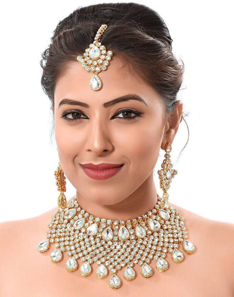 May include: A gold and white bridal jewellery set with a necklace, earrings, and a tikka. The necklace features a large, teardrop-shaped pendant with smaller stones surrounding it. The earrings are also teardrop-shaped and have a similar design to the necklace.