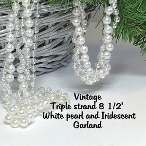 May include: Vintage triple strand 8.5-foot white pearl and iridescent garland. The garland features large, round white pearls and smaller, clear iridescent beads. The garland is draped in front of a gray wicker basket and evergreen branches.