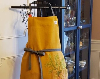 Op de afbeelding: Een mosterdgele linnen schort met een grijze strikband en een grote zak aan de voorkant. De zak heeft een geborduurd ontwerp van een bos wortels en groene bladeren.