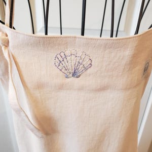以下が含まれることがあります： 薄いピンク色のリネンエプロン。青と白の糸で貝殻の刺繍が施されています。エプロンにはポケットと調節可能なストラップが付いています。貝殻のデザインはエプロンの胸の中央に配置されています。