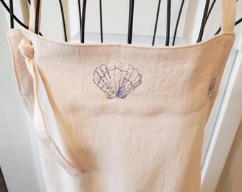 May include: A light pink linen apron with a seashell embroidery in blue and white thread. The apron has a pocket and adjustable straps. The seashell design is centered on the chest area of the apron.